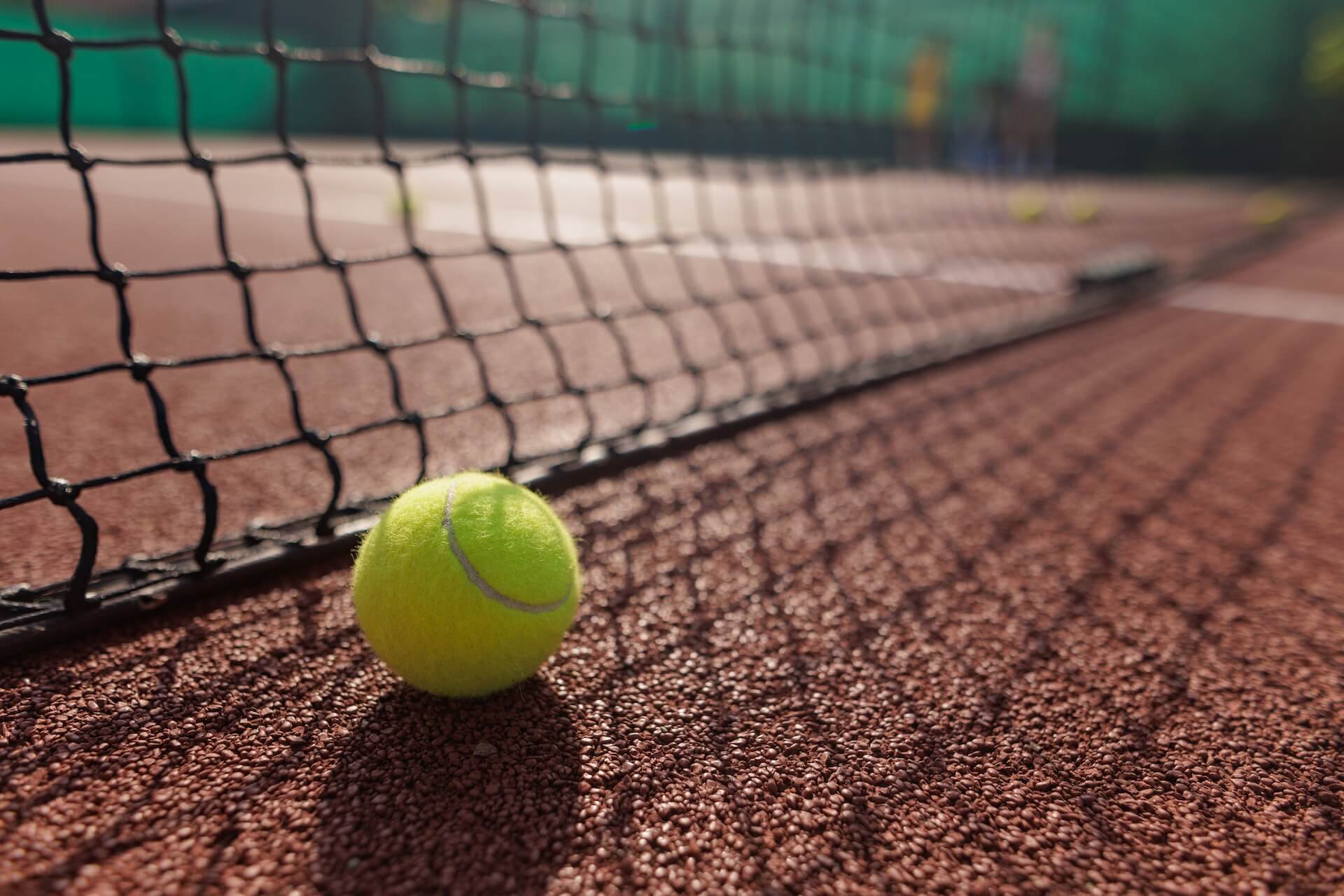best-tennis-court-surface-00 Zoomed in photo of a tennis ball next to a tennis net on a tennis court | Featured image for the Choosing the Best Tennis Court Surface blog page.